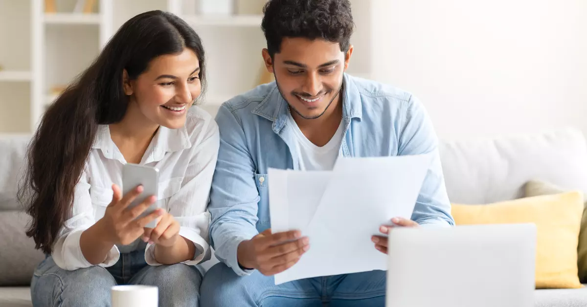 A smiling couple sitting together, reviewing documents and using a smartphone