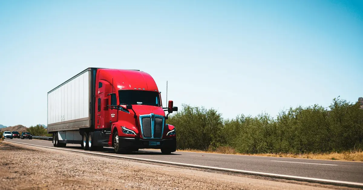 Red commercial semi-truck driving on a highway under a clear blue sky, with cars following behind