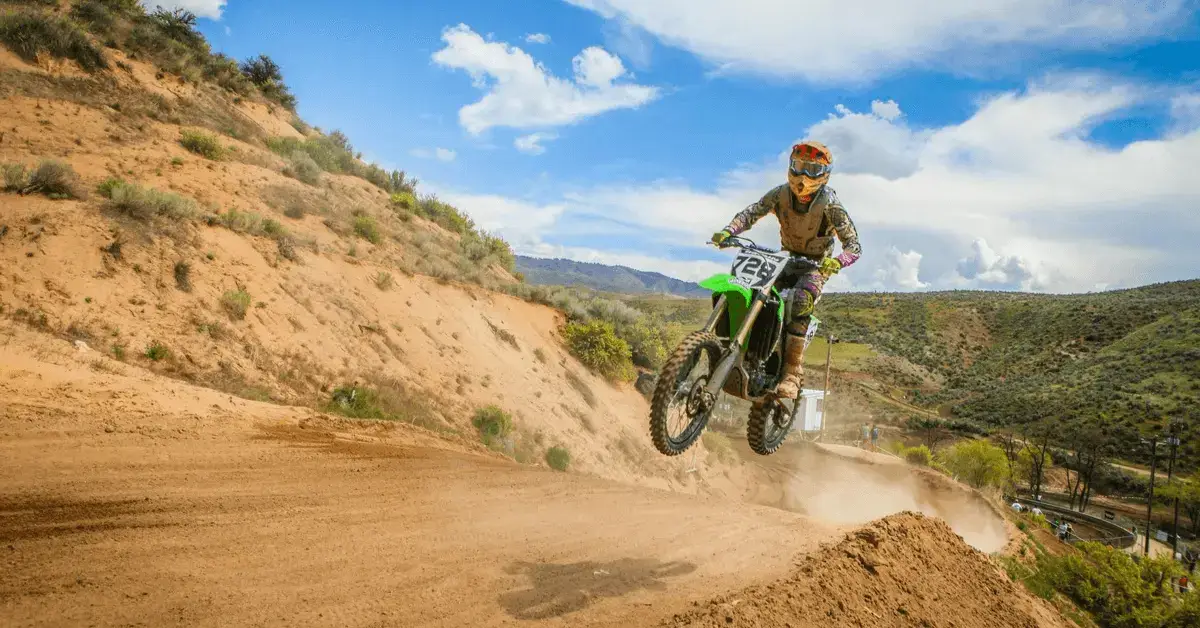 Person riding an ATV on a dirt track, wearing a helmet and protective gear, with a desert landscape and hills in the background