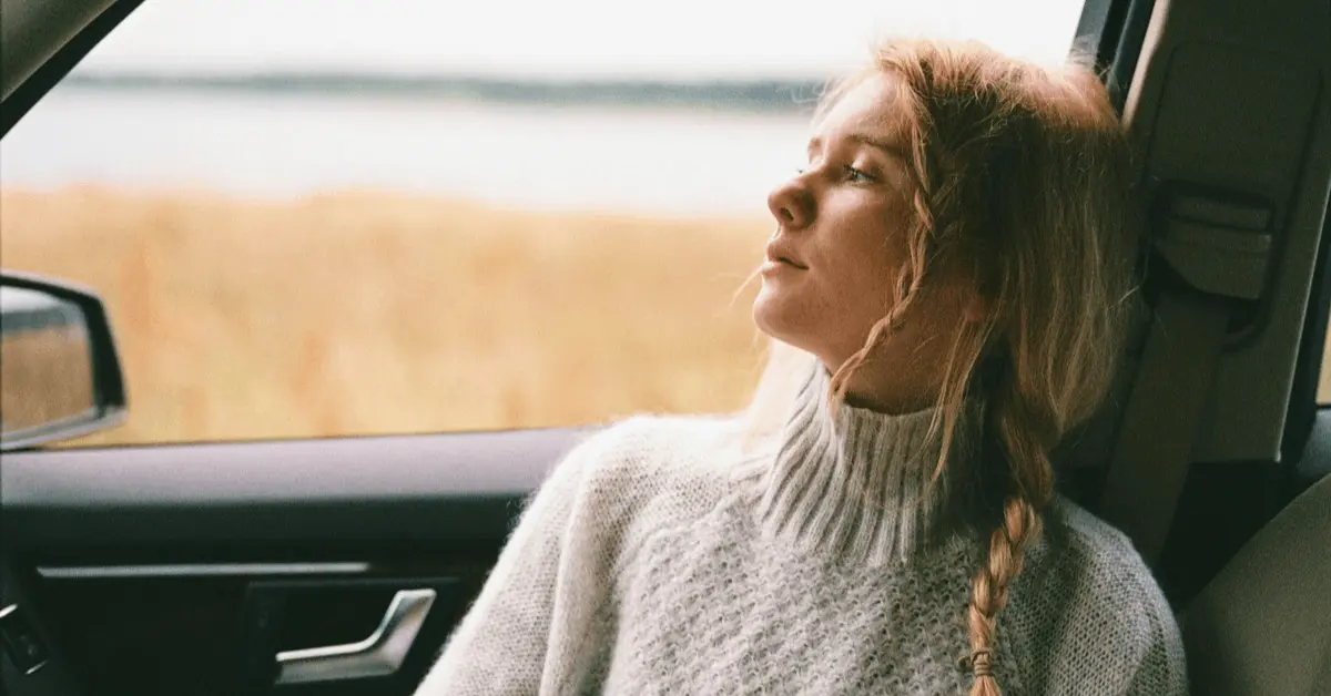 A woman is seated in the passenger side of a car, looking out the window at a serene field, reflecting on her thoughts or mental health in a moment of tranquility.