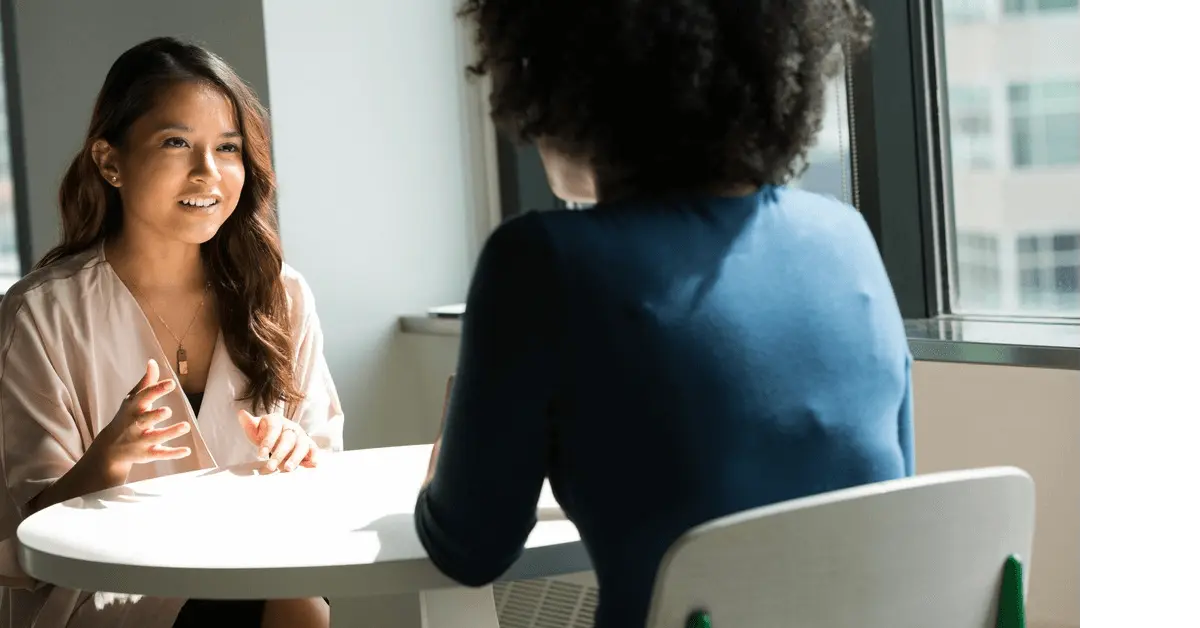 Two women sitting at a table in a discussion, with one explaining her situation