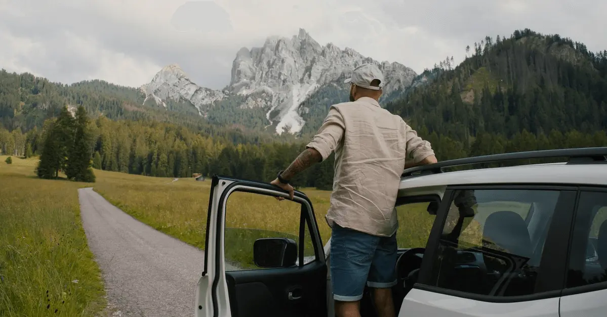 Man standing outside his car on a mountain road, admiring a scenic view of a meadow and distant rocky peaks