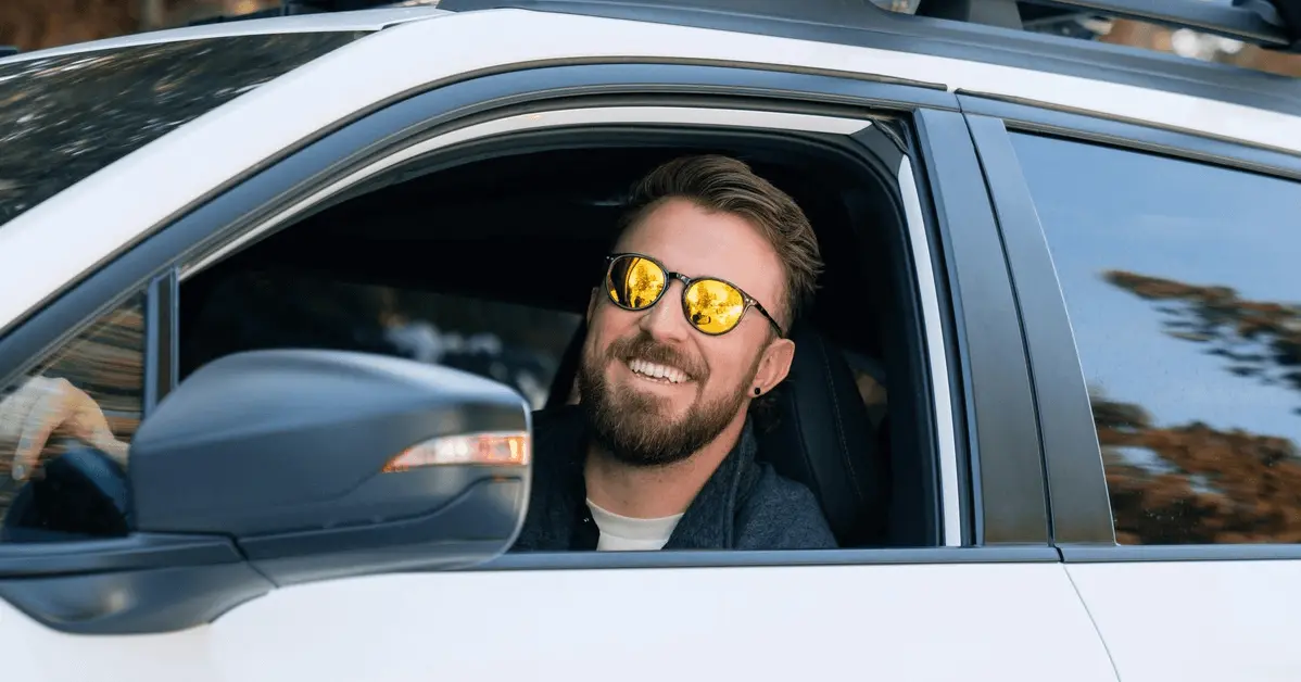 Smiling man wearing sunglasses, sitting in the driver's seat of a white car with the window down