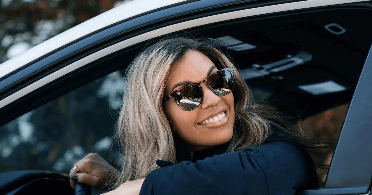 Smiling woman wearing sunglasses, sitting in the driver's seat of a car, looking out the window with her hand on the steering wheel