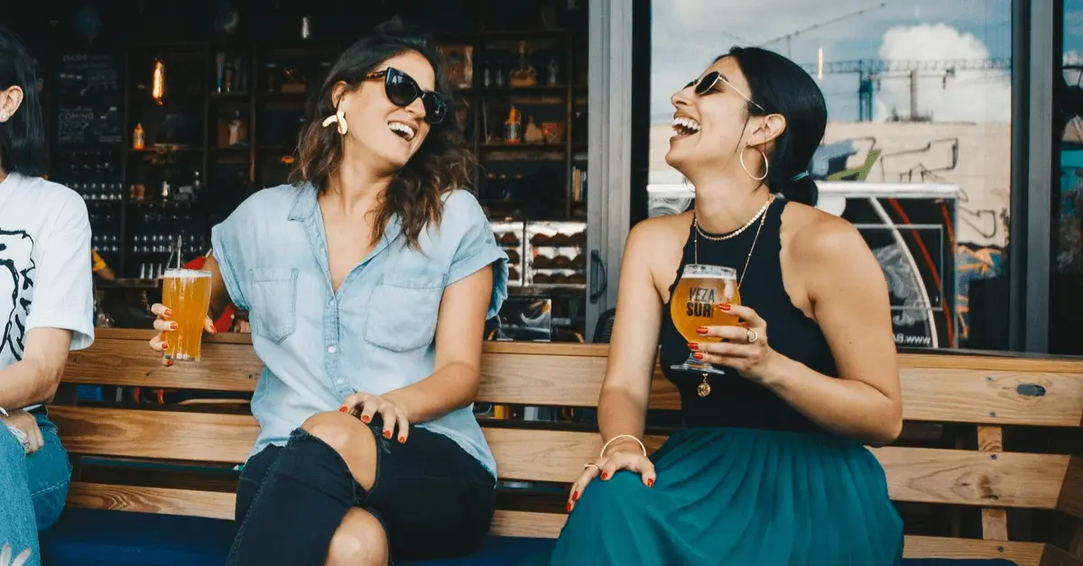 Two women sitting outdoors, laughing with a glass of beer in their hands