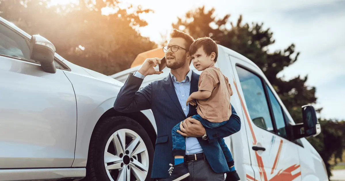 Father holding his young son while talking on the phone beside a car and tow truck