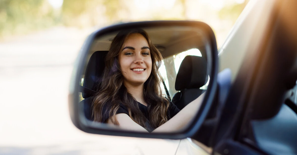 A young woman smiling as she sits in the driver's seat, reflected in the car's side mirror