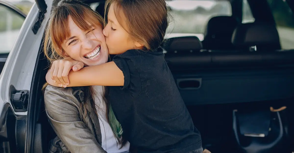 A smiling woman sitting at the open trunk of a car, receiving a kiss on the cheek from her daughter