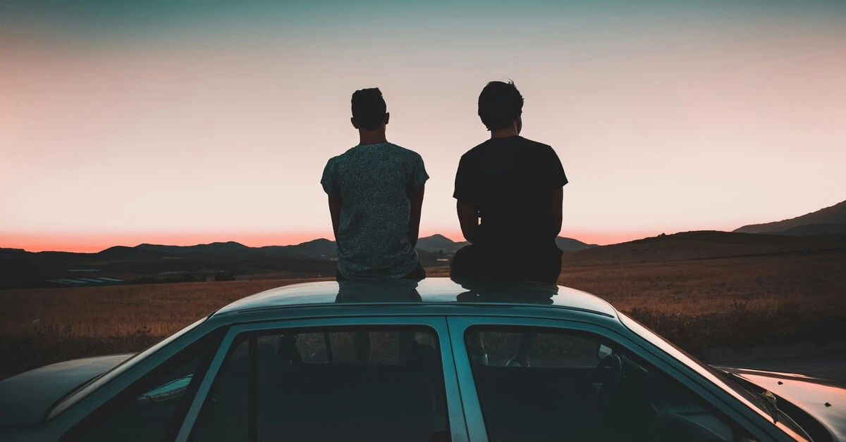 Two people sitting on the roof of a car at sunset, overlooking an open field and distant rolling hills