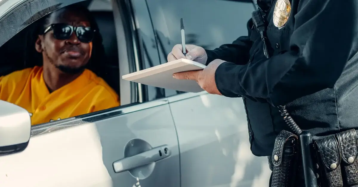 Police officer writing a ticket while speaking to a driver seated in a car during a traffic stop