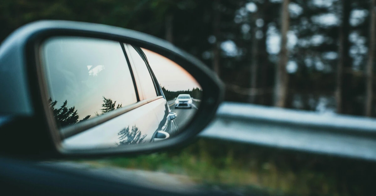 View of a car in a side mirror on a forest-lined road