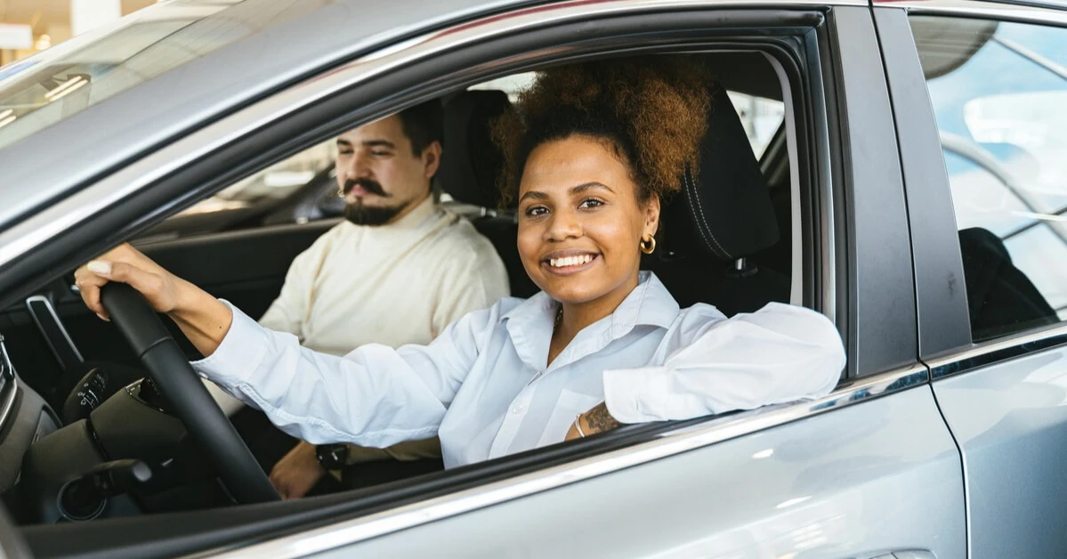 Smiling woman sitting in the driver’s seat of a gray sedan with a male passenger next to her