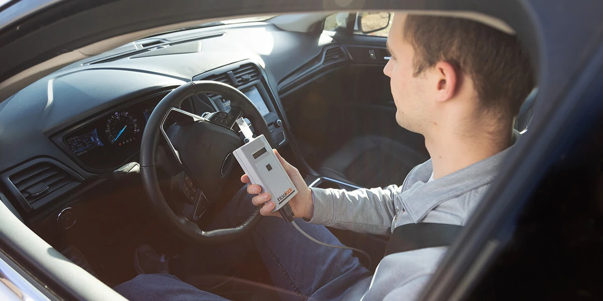 A man sitting in the driver's seat of a car, holding an Intoxalock ignition interlock device, preparing to take a breath test before starting the vehicle.