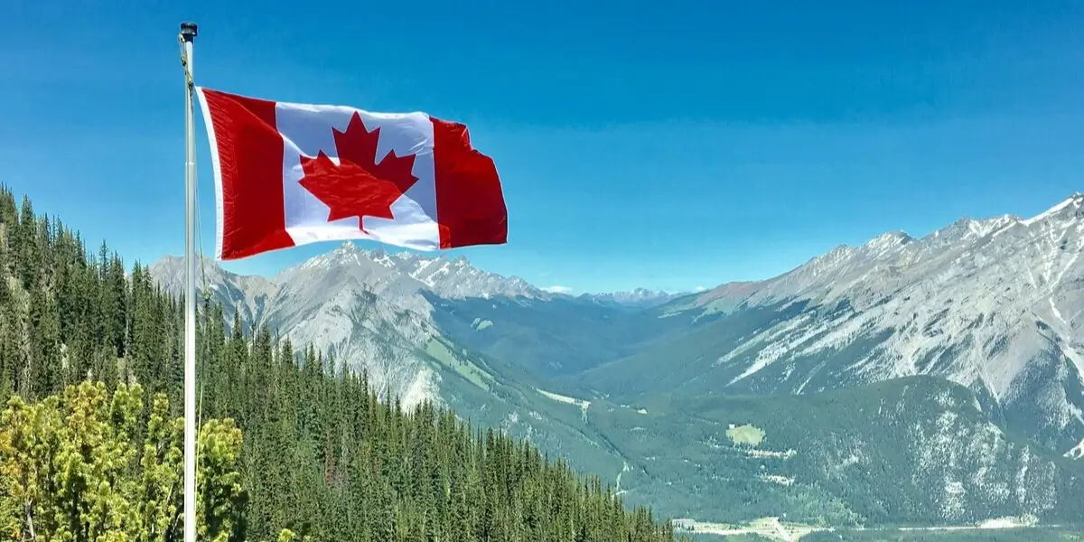 A Canadian flag waving on a flagpole against a backdrop of mountains and evergreen forests under a clear blue sky