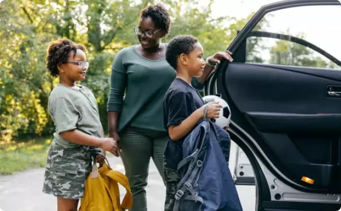 A smiling woman stands beside two children holding backpacks and a soccer ball near an open car door